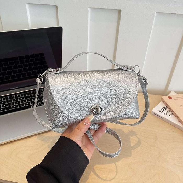 A silver pebbled leather handbag with a top handle and shoulder strap, held by a hand over a light wood desk with a laptop and books in the background.