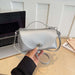 A silver pebbled leather handbag with a top handle and shoulder strap, held by a hand over a light wood desk with a laptop and books in the background.