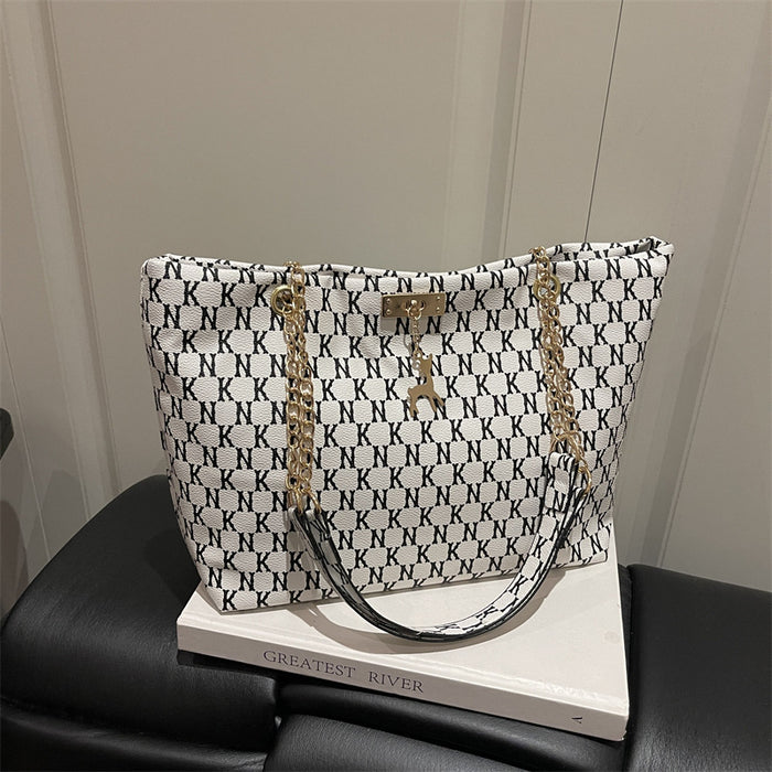 A white tote bag with a black repeating monogram pattern and gold-tone chain handles, displayed on a white book resting on a black leather chair.
