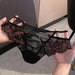 Black lace and mesh panties held up by two hands against an indoor background with gray flooring and light wood paneling.