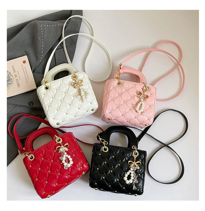 Four quilted mini handbags in white, pink, red, and black, arranged on a light beige surface with a gray jacket and a small book in the background. Each bag has a top handle, a long shoulder strap, a diamond-quilted pattern, gold-tone hardware, and a pearl tassel charm.