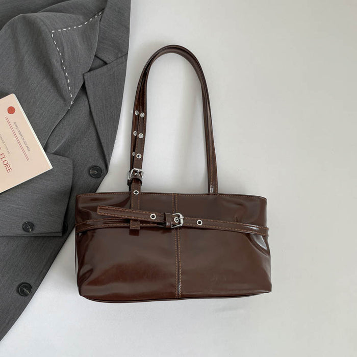 A glossy dark brown leather shoulder bag with silver-tone hardware, displayed on a light gray surface next to a gray blazer and a small card.