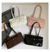 Four structured shoulder handbags arranged on a light neutral background: one cream, one blush pink, one dark brown patent leather, and one black patent leather. A dark gray fabric bag and a small paper tag are partially visible on the left.