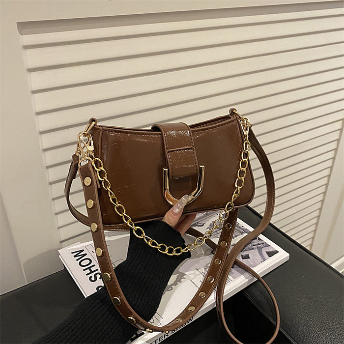 A brown faux-leather shoulder bag with a gold-tone chain strap and gold studs, held by a hand over a magazine on a black table.