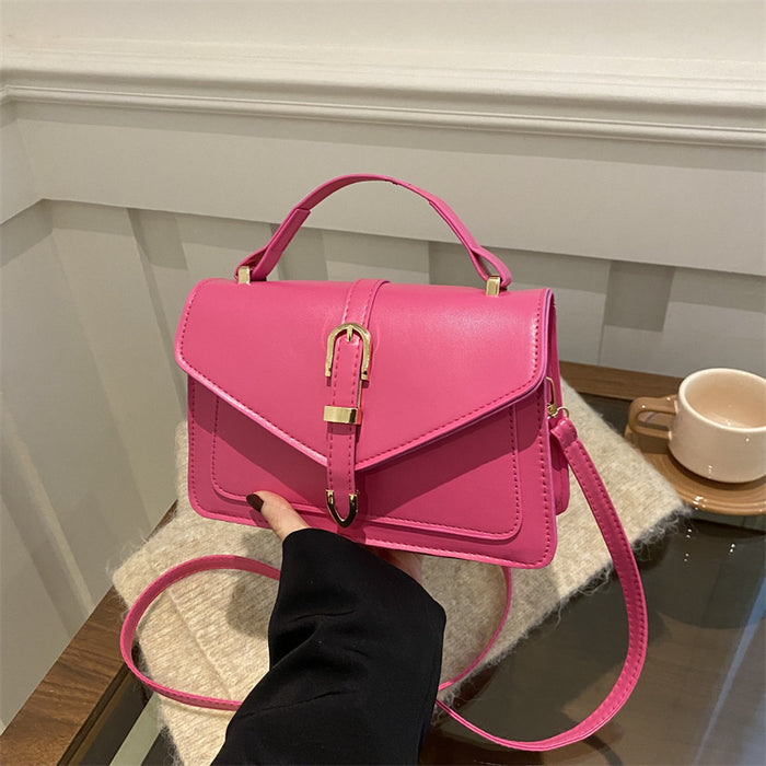 A bright pink structured handbag with a top handle, flap front, and gold-tone clasp, held by a hand over a cream knit blanket in an indoor setting.