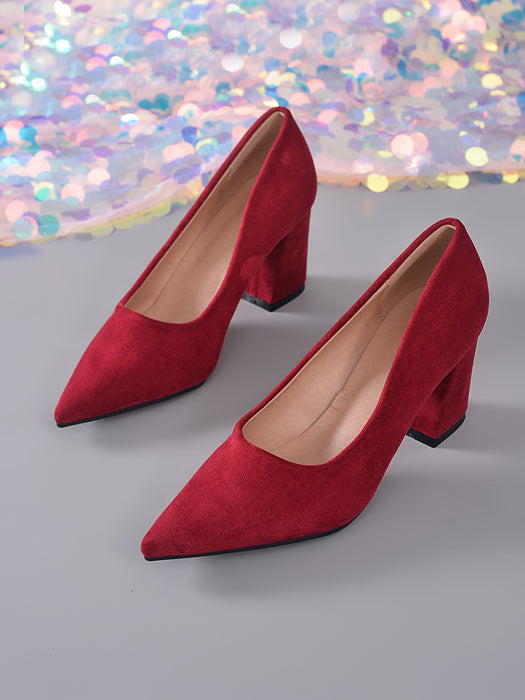 A pair of red suede high-heeled pumps with pointed toes and block heels, displayed on a gray surface with a colorful sequined fabric backdrop.