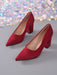 A pair of red suede high-heeled pumps with pointed toes and block heels, displayed on a gray surface with a colorful sequined fabric backdrop.