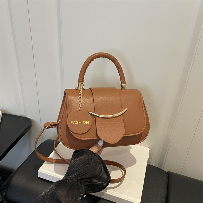 A medium brown faux-leather handbag with a top handle, a round hanging charm that reads 'FASHION', and a curved gold-tone metal accent on the front, shown resting on a white box with a black sleeve holding it.