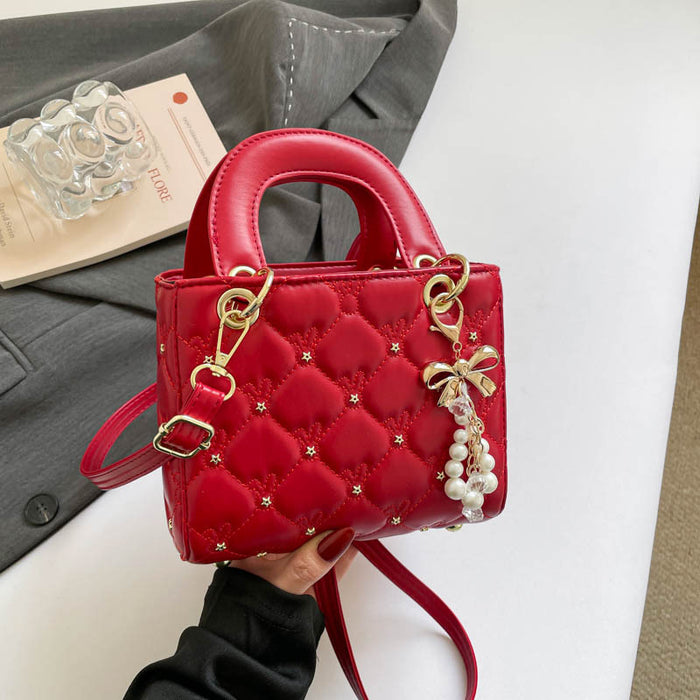 A red quilted top-handle handbag with gold-tone hardware, pearl charm details, and a shoulder strap, held by a hand against a styled background with a gray blazer, book, and clear glass object.