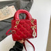 A red quilted top-handle handbag with gold-tone hardware, pearl charm details, and a shoulder strap, held by a hand against a styled background with a gray blazer, book, and clear glass object.