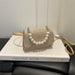 A small woven beige handbag with a curved flap adorned with white faux pearls and silver-tone hardware, resting on a white book with a black leather chair behind it.
