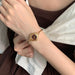 A close-up of a person's wrist wearing a slim gold-tone watch with a round dark dial and matching gold bracelet, styled with a brown top and white draped fabric.