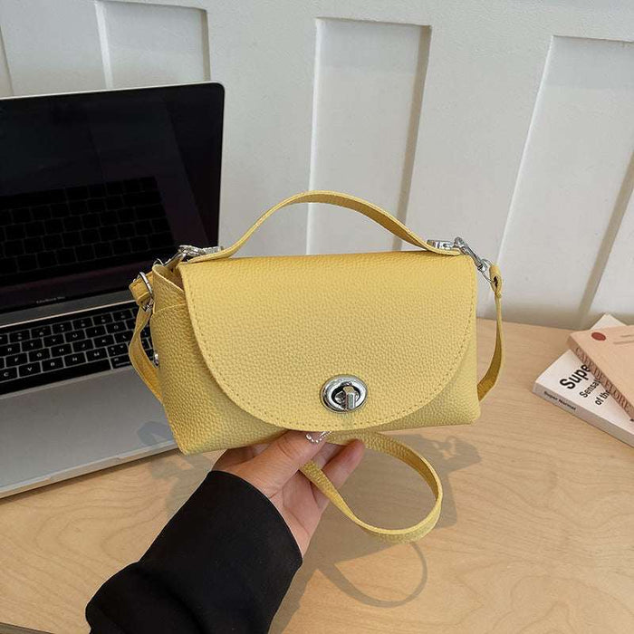 A light yellow pebbled leather handbag with a top handle and silver-tone hardware, held by a hand over a light wood desk with a laptop and book in the background.