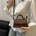 A person wearing a white blouse and light blue jeans carries a small brown top-handle crossbody bag with a matching shoulder strap. The bag has a structured rectangular shape, a top handle, a front flap, and a zipper closure.
