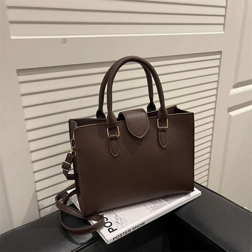 A dark brown textured leather handbag with double top handles and a detachable shoulder strap, resting on a black surface next to a white magazine in front of a white slatted door.