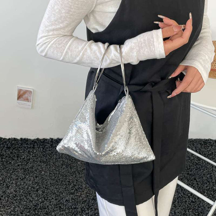 A person wearing a white long-sleeve top and a black apron-style garment holds a silver sequined slouchy shoulder bag with thin metallic handles.
