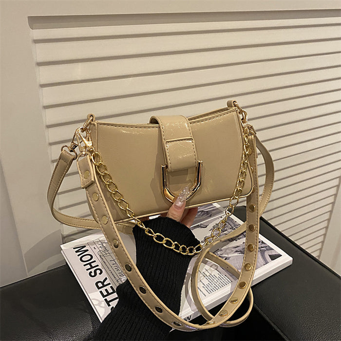A beige faux-leather shoulder bag with a gold chain strap and gold-tone hardware, held by a hand over a magazine on a black surface in front of a white slatted wall.
