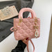 A pink quilted top-handle handbag with gold-tone hardware, pearl charm, and rhinestone details, held by a hand over a white surface with a gray jacket and small card in the background.