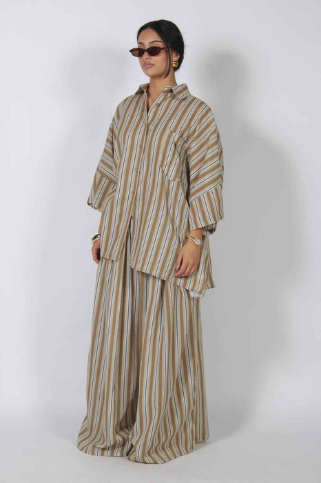 A woman modeling a matching two-piece striped lounge set consisting of an oversized long-sleeve shirt and wide-leg pants, in beige, brown, and white vertical stripes, set against a plain light gray studio background.
