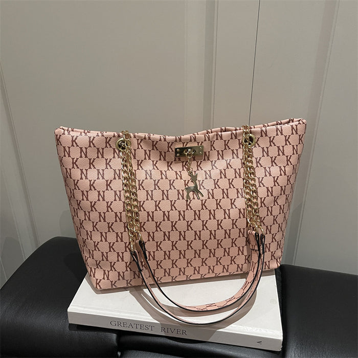 A pink monogram tote bag with gold-tone chain straps and a small deer charm, displayed on a white book resting on a black leather surface against a light paneled wall.