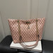A pink monogram tote bag with gold-tone chain straps and a small deer charm, displayed on a white book resting on a black leather surface against a light paneled wall.