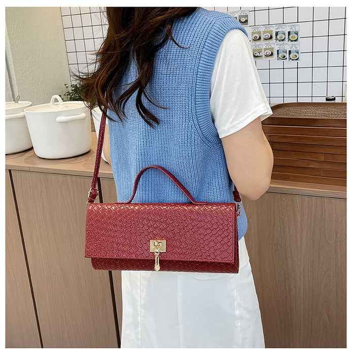 A woman wearing a blue knit vest over a white t-shirt and white skirt carries a red textured crocodile-embossed handbag with a gold clasp in a kitchen setting.