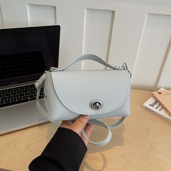 A light blue pebbled leather handbag with a top handle and shoulder strap, held by a hand over a wooden desk with a laptop in the background.