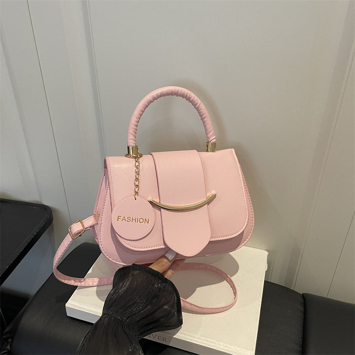 A light pink faux leather handbag with a top handle, a circular 'FASHION' tag, and a gold-tone curved accent on the front, resting on a white box with a black sleeve holding the strap.