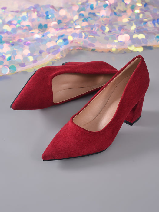 A pair of red suede pointed-toe pumps with block heels, displayed on a gray surface with a decorative iridescent sequin fabric in the background.