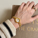 Hand wearing a gold watch with a textured sleeve on a beige surface