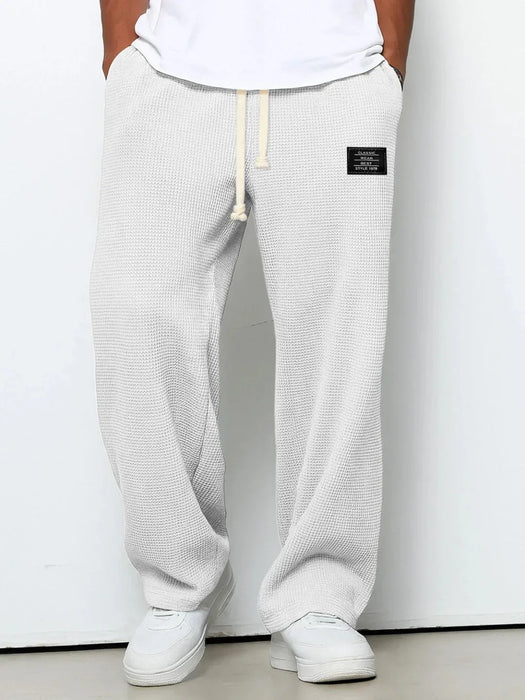 Person wearing white textured sweatpants with a small black logo on a plain background