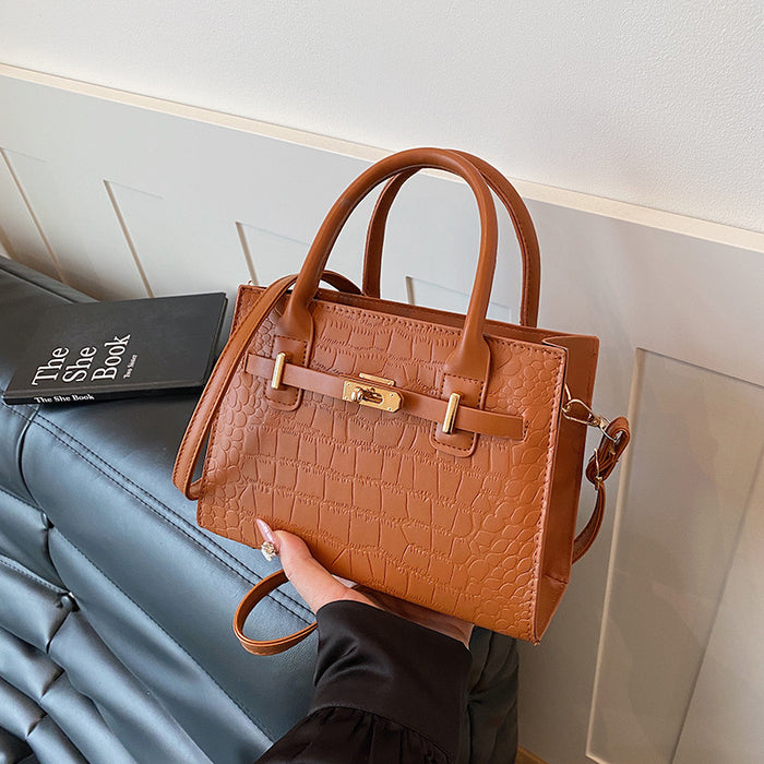 A tan crocodile-embossed leather handbag with gold-tone hardware, held by a hand over a dark gray leather sofa next to a book.