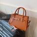 A tan crocodile-embossed leather handbag with gold-tone hardware, held by a hand over a dark gray leather sofa next to a book.