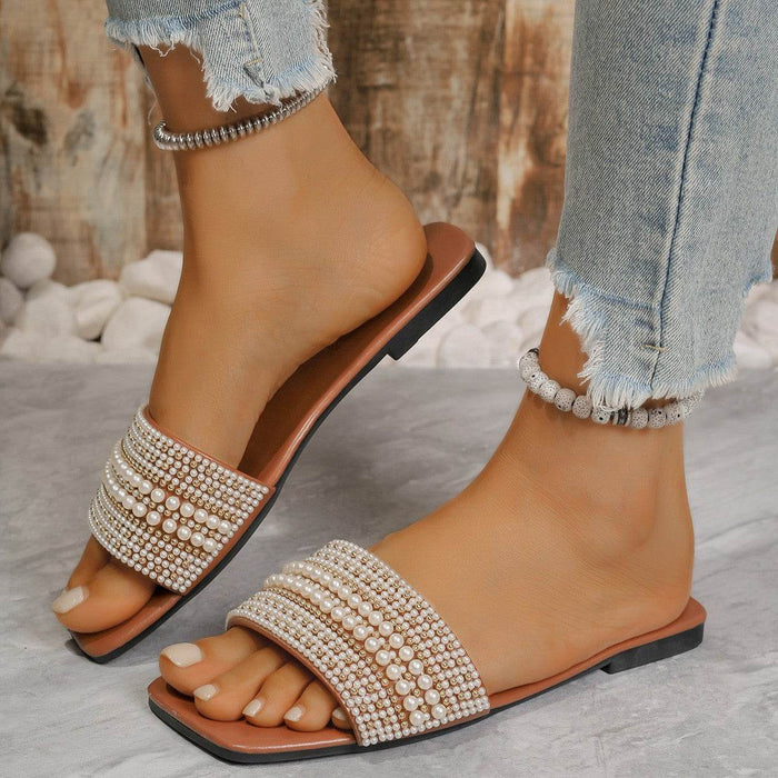 Tan sandals with pearl embellishments worn by a person with denim pants against a natural stone background.