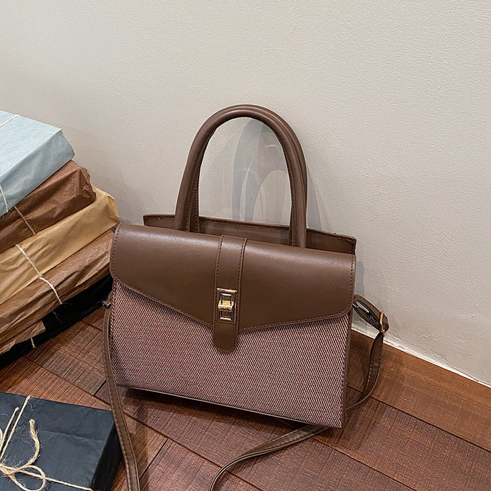 A structured handbag with a brown faux-leather top and flap, a textured dusty pink woven lower body, and a gold-tone front clasp, shown on a wooden floor next to stacked gift boxes.