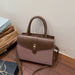 A structured handbag with a brown faux-leather top and flap, a textured dusty pink woven lower body, and a gold-tone front clasp, shown on a wooden floor next to stacked gift boxes.