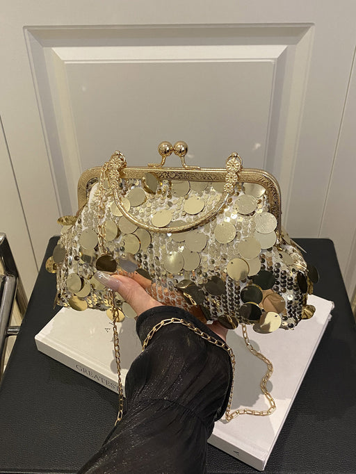 A gold-tone kiss-lock handbag covered in round metallic coin embellishments and hanging clear beads, held by a person wearing a black sleeveless top. The bag is displayed on a white book on a black table in front of a white paneled wall.