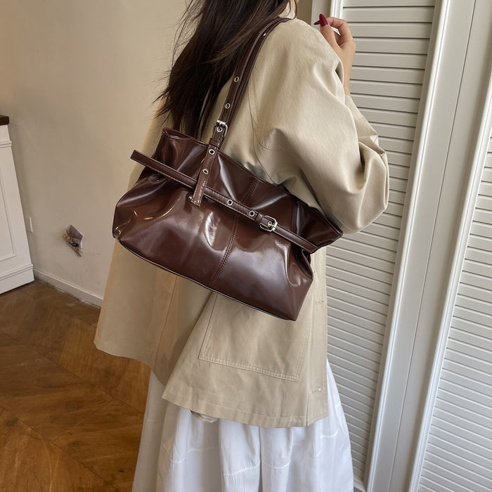 A person wearing a beige oversized shirt and white skirt carries a shiny dark brown shoulder bag with silver hardware in an indoor room with beige walls and a slatted door.