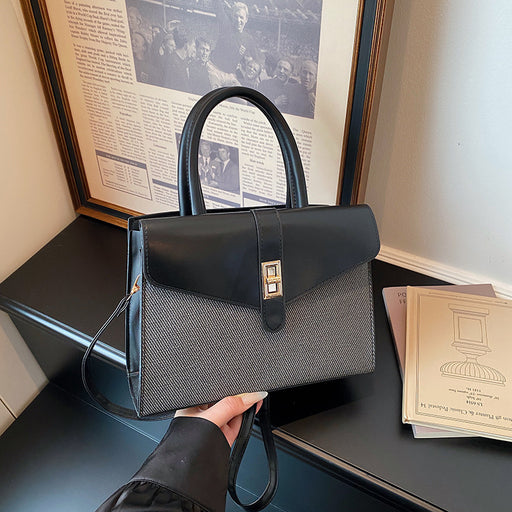 A structured black handbag with a textured gray woven lower body and a gold-tone front clasp, held by a hand in a black sleeve. The bag is positioned on a dark table with a framed newspaper print and a book in the background.