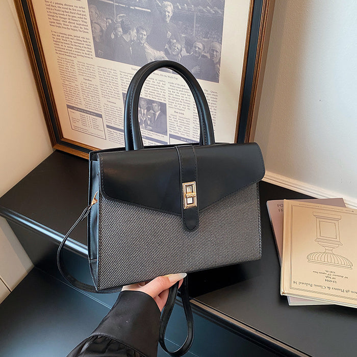 A structured black handbag with a textured gray woven lower body and a gold-tone front clasp, held by a hand in a black sleeve. The bag is positioned on a dark table with a framed newspaper print and a book in the background.