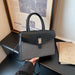 A structured black handbag with a textured gray woven lower body and a gold-tone front clasp, held by a hand in a black sleeve. The bag is positioned on a dark table with a framed newspaper print and a book in the background.