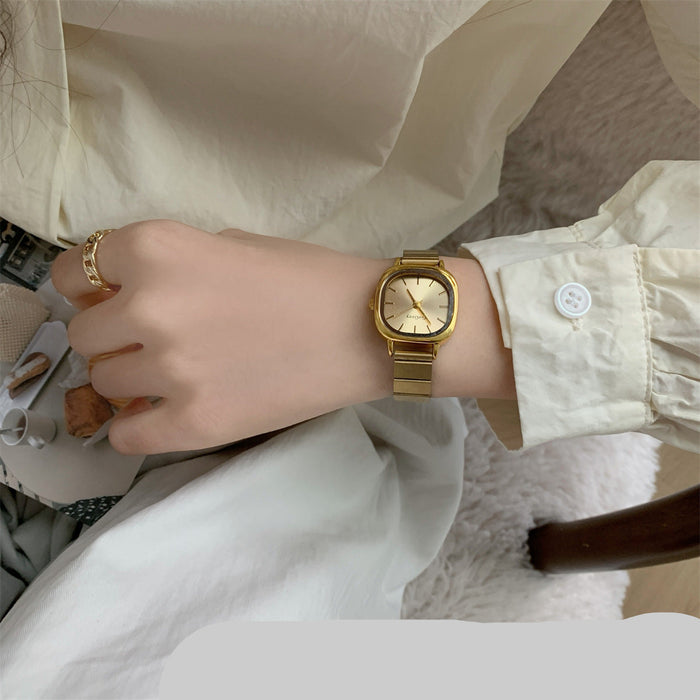 A gold-tone metal link watch with a round dial worn on a person's wrist, shown in a close-up lifestyle shot with a cream-colored shirt and soft neutral background.