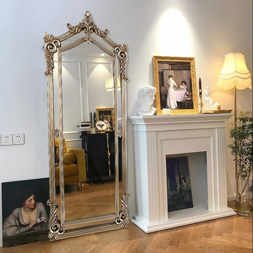 Decorative mirror with ornate frame next to a fireplace with a painting and lamp.