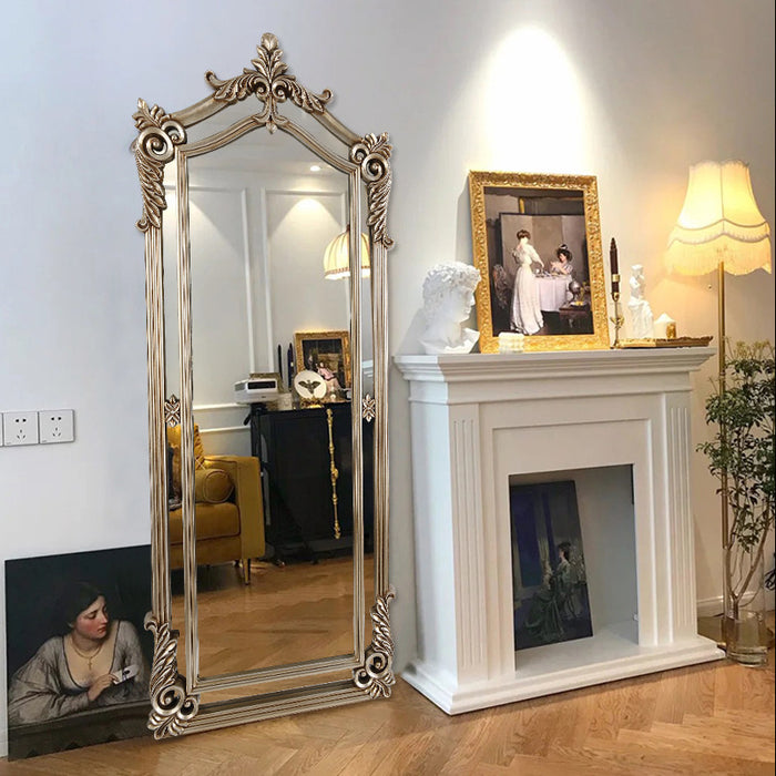 Decorative mirror with ornate frame next to a fireplace with a painting and lamp.