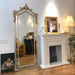 Decorative mirror with ornate frame next to a fireplace with a painting and lamp.