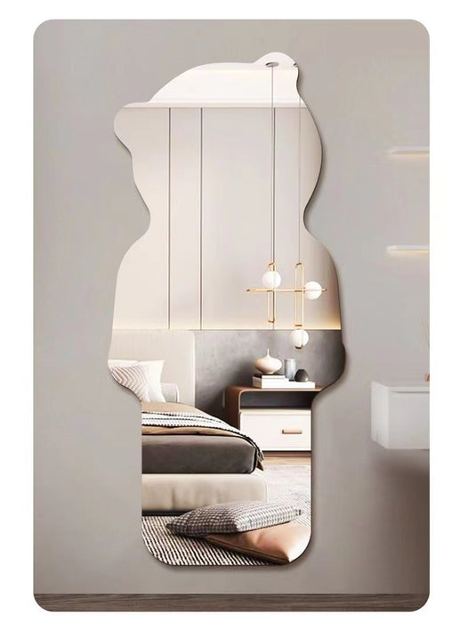 An irregularly shaped wall mirror with a wavy silhouette, mounted on a light gray wall. The mirror reflects a modern bedroom interior with a bed, bedside table, pendant lights, and neutral-toned decor.