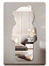 An irregularly shaped wall mirror with a wavy silhouette, mounted on a light gray wall. The mirror reflects a modern bedroom interior with a bed, bedside table, pendant lights, and neutral-toned decor.