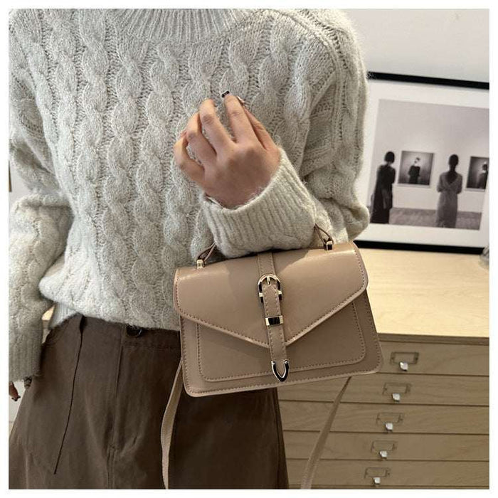 A person wearing a cream cable-knit sweater and brown pants carries a beige structured leather shoulder bag with a flap front and silver-tone buckle.
