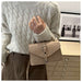 A person wearing a cream cable-knit sweater and brown pants carries a beige structured leather shoulder bag with a flap front and silver-tone buckle.