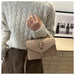 A person wearing a cream cable-knit sweater and brown pants carries a beige structured leather shoulder bag with a flap front and silver-tone buckle.